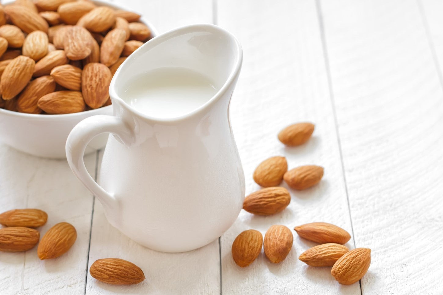 Creamy almond milk in a white jug with whole almonds on table.