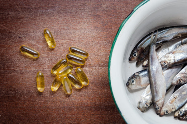 Fish oil capsules next to a bowl of fresh fish on a wooden surface.
