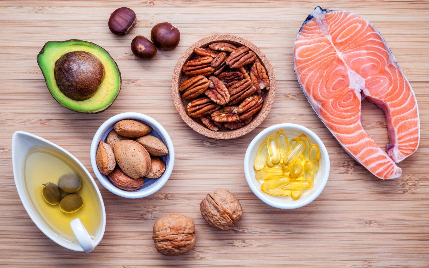 Assorted omega-rich foods including salmon, avocado, nuts, and oil capsules on a wooden background.