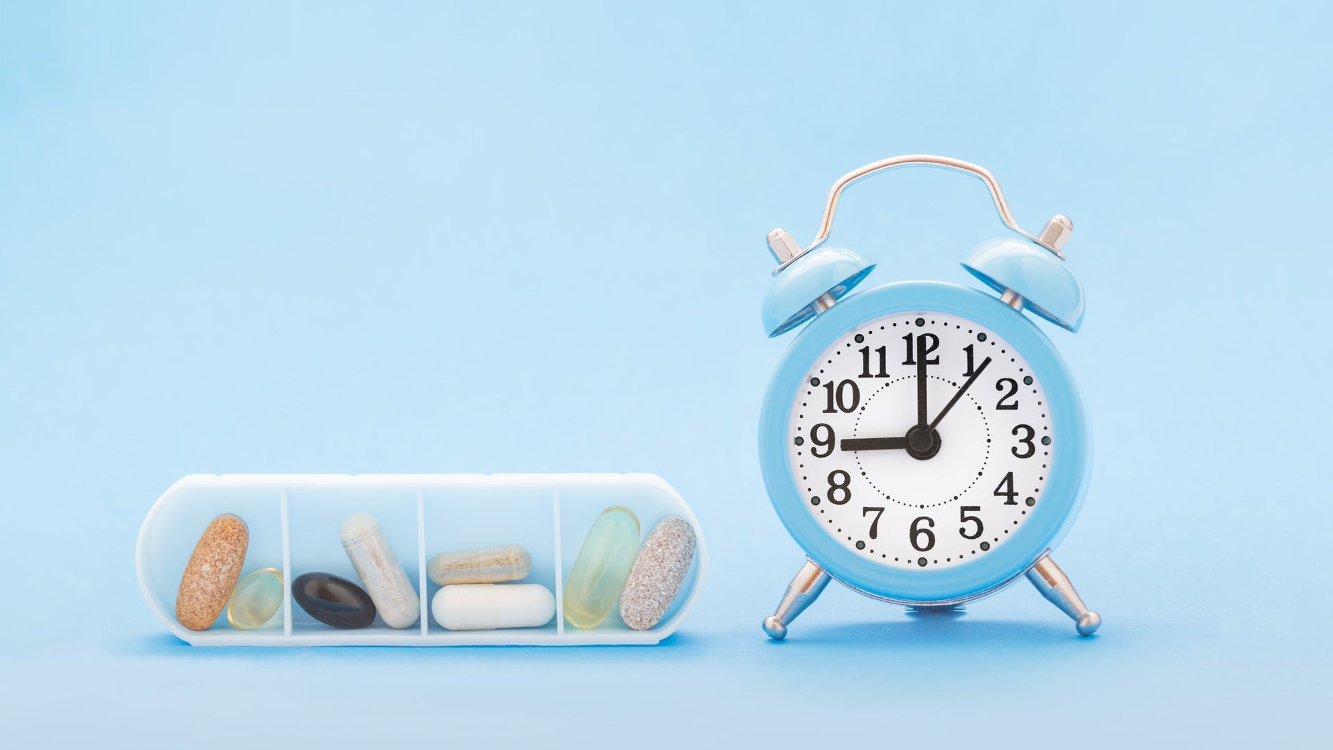 Pill organizer and blue alarm clock on light blue background.